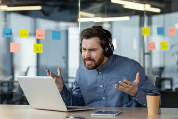 Frustrated man wearing headphones during remote work video conference in modern office