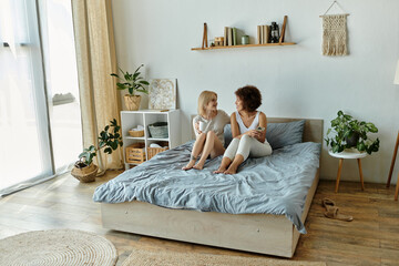 Two women in pajamas are sitting on a bed, chatting and enjoying their morning.