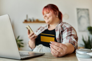 A woman holds a credit card and a phone while smiling at a laptop.