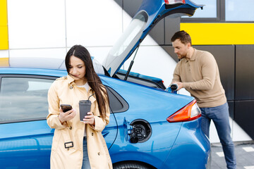 Young couple man and woman traveling by electric car having stop at charging station. Boyfriend...