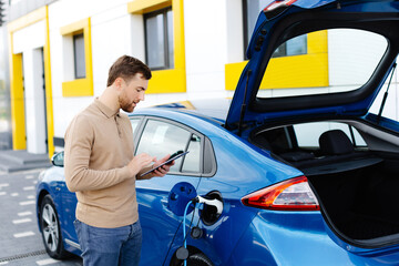 A young man is charging an electric car while standing at a station and holding a tablet