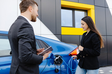 Sales manager showing how to charge electric car to a young client woman, plugging wire into the...