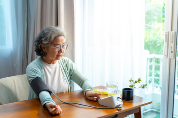 Asian senior mature woman patient with hypertension using digital blood pressure monitor measuring blood pressure by herself in bedroom at home. Elderly people self-care health check-up concept.