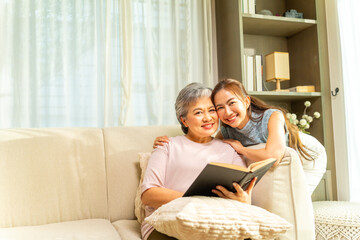 Asian senior mature woman and adult daughter spending time together reading a book on sofa in living room. Happy family relax and enjoy indoor lifestyle with leisure activity together at home.