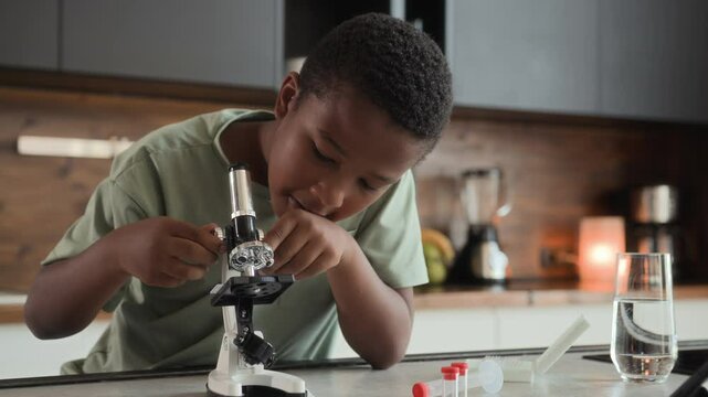 African American boy is sitting at table in kitchen, studying and doing homework. He uses microscope, discovering knowledges during homeschool. Black skinned schoolboy is interested and curious