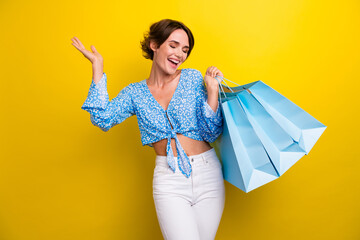 Portrait of overjoyed nice girl with short hair wear blue blouse hold shopping bags raising palm up...