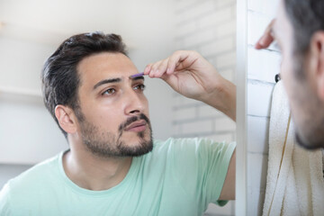 Fototapeta premium Handsome man grooming in front of the mirror in the bathroom. Young guy depilates with tweezers 