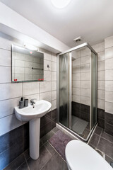 Standard modern bathroom featuring a corner shower cabin, sink, mirror, and light. The space is adorned with dark grey tiles, creating a sleek and contemporary look.