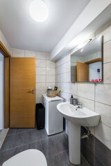 Standard bathroom interior featuring a white pedestal sink, a wall-mounted mirror, and a washing machine positioned in the corner. Space is functional and neatly arranged, showcasing modern fixtures 
