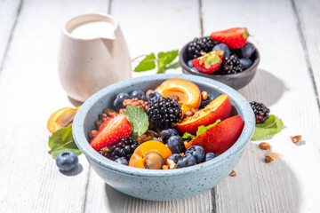 Granola breakfast with fruit and berry, summer whole grain tasty breakfast bowl with a lot of fruit and berries, with milk jug on white wooden table
