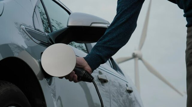 Unrecognizable person disconnects charging cable from an electric car charging alongside wind turbine, symbolizing sustainable energy and green technology. Male hands unplug power connector from EV
