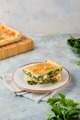 A rectangular cut Greek cake with spinach and cheese on a plate on a gray concrete background. Spinach recipes.