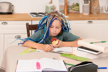 At the kitchen table, a young child with colorful braids focuses on homework, eating an apple while...