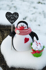A toy penguin with a red and white scarf stands in the snow and sells snowballs