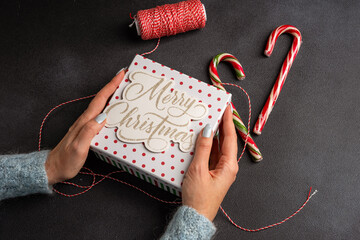 Christmas composition on a dark background: Beautiful female hands holding a gift liht box, A spool of red and white thread and two candy canes.