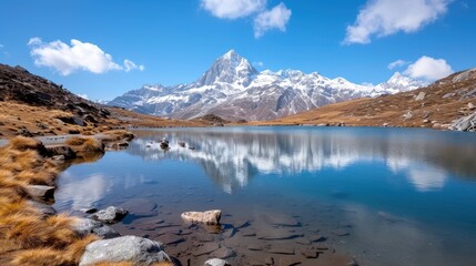 Naklejka premium A peaceful alpine lake provides a breathtaking view of a snow-covered peak reflected in its calm waters, set amid a barren landscape with sparse vegetation and clear skies.