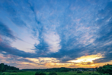 Obraz premium A man stands on a bridge, taking a photo of a stunning sunset with a cellphone. The vibrant colors of the sky and the lush green foliage create a picturesque scene.
