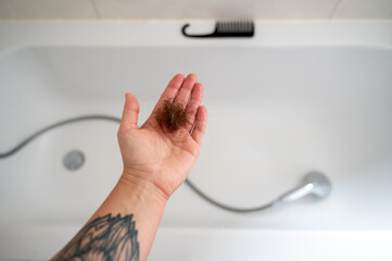 Hand holding clump of fallen hair on palm after washing hair. In background, bathtub, shower, and...