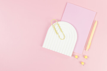 Top view of pink notebook, yellow pen, paperclip, pins, white arch tray on pink background. School, office wallpaper. Flat lay, copy space.