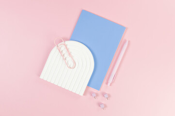Top view of blue notebook, pink pen, paperclip, pins, white arch tray on pink background. School,...