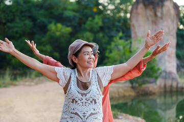 Two elderly Asian women, one in orange dress, other in white lace top, standing on dirt path with green trees, arms raised, smiling. Celebrating freedom and friendship, enjoying outdoor activity