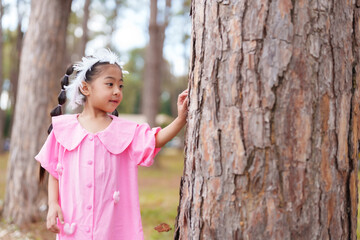 Asian girl in pink dress standing near tree in park. Looking to side, enjoying natural environment. Concept of childhood wonder, exploration, and playful moments in a sunlit forest setting.