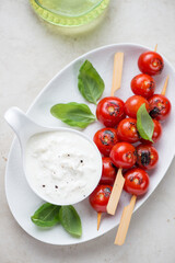 White plate with fresh stracciatella di bufala, roasted cherry tomato skewers and basil, vertical shot on a light-beige stone surface, high angle view