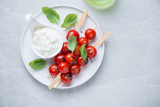 Skewers with roasted cherry tomatoes, stracciatella cheese and basil on a white plate, above view on a light-grey granite background, horizontal shot with space