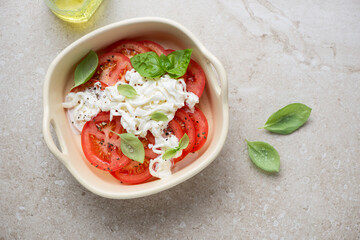 Serving tray with tomato slices, basil and stracciatella or stretched curd cheese, top view on a beige stone background, horizontal shot
