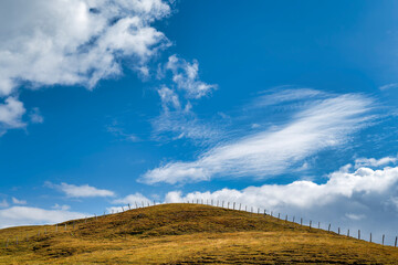 Pasture fence on an autumnal grassy hill in the mountains against a magnificent blue sky. Big clouds are gathering around.