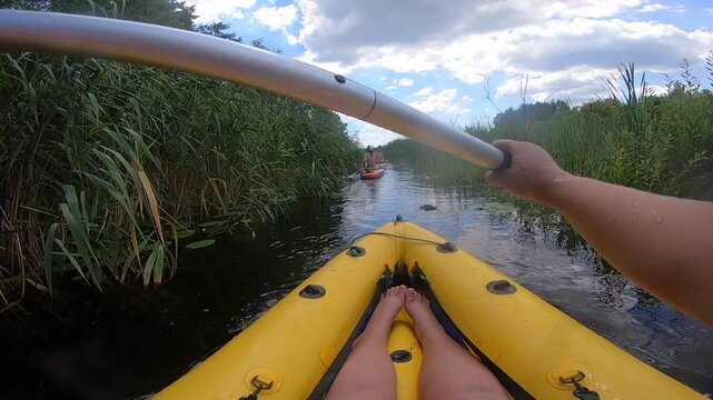 Person floating in inflatable boat along river through thickets of green reeds on sunny summer day. People kayaking along river. Man in boat rowing oars. Sport, action, activity, lifestyle. POV