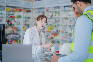 Obraz premium A female pharmacist is talking with a construction worker who needs medicine for pain and discomfort. Pharmacist in a pharmacy with medicines on shelves