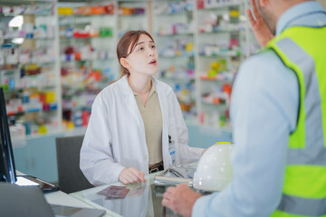 Obraz premium A female pharmacist is talking with a construction worker who needs medicine for pain and discomfort. Pharmacist in a pharmacy with medicines on shelves