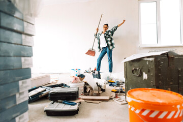 Young smiling woman with sweeping brush is dancing in renovated room. Break between cleaning of...