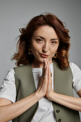 A stylish Asian woman in a green vest with a soft smile stands against a gray background, her hands clasped in prayer.