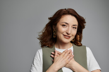 A stylish Asian woman in a vest smiles warmly, hands clasped over her heart, against a simple gray background.