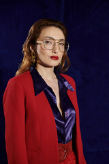 A stylish Asian woman in a red blazer and silk shirt poses confidently against a dark background.