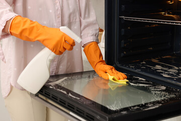 Woman cleaning oven door with rag and detergent in kitchen, closeup