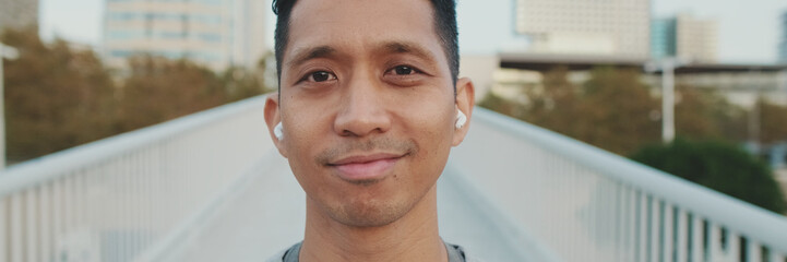 Close up, athlete in wireless headphones, a guy in sportswear, stands on bridge and looks at camera, listens to music, panorama