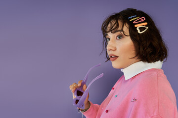 A young woman in a pink sweater gazes off-camera, holding purple sunglasses.