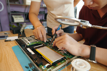 Repairman soldering board of laptop under magnifier glass together with his colleague in workshop