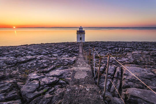 Path crossing the limestone burren leading to a white lighthouse with the sea and setting sun in the background; Blackhead, County Clare, Ireland