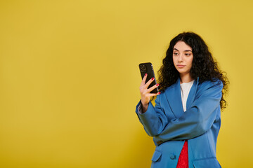 Naklejka premium Young brunette woman with curly hair, wearing a blue jacket, looking at her cell phone in a studio with a yellow background.