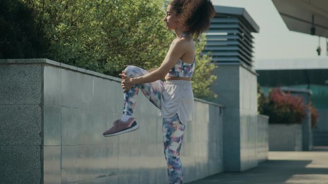 Young curly-haired girl pulling knee up to chest, then placing one leg on wall and doing hamstring stretch before outdoor training in city