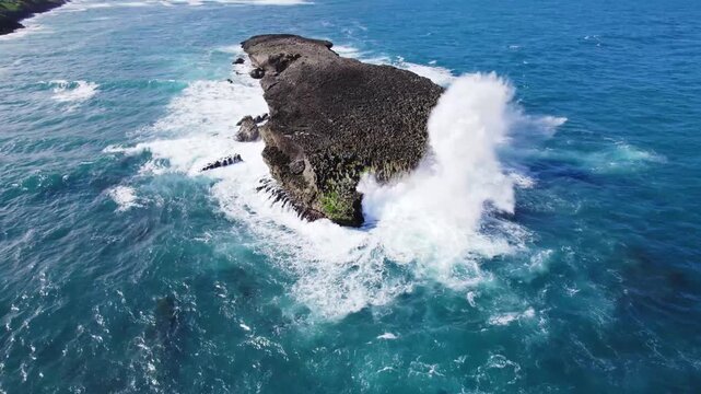 An aerial shot of a rugged islet being pummeled by powerful ocean waves. The scene captures the raw beauty and intensity of nature as the turquoise water contrasts with the rocky outcrop.