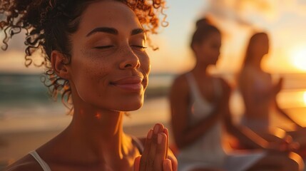 A woman meditates on the beach with two other women in the background, bathed in the warm glow of the setting sun.