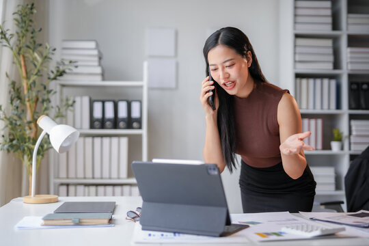 Businesswoman is having a heated conversation on her smartphone, leaning on her desk and looking frustrated and angry
