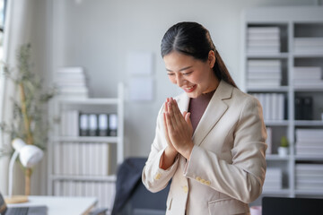 Fototapeta premium Young professional woman is standing in her office, smiling and greeting someone with her hands pressed together in a respectful gesture