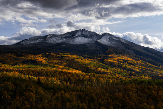 Scenic vista of fall colored forest on the slopes in front of snow-capped mountain peaks under grey clouds and a blue sky in the distance; Colorado, United States of America