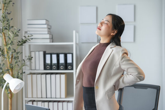 Young manager woman holding her lower back while standing up from her desk in her office. She is relieving back pain after many hours of work